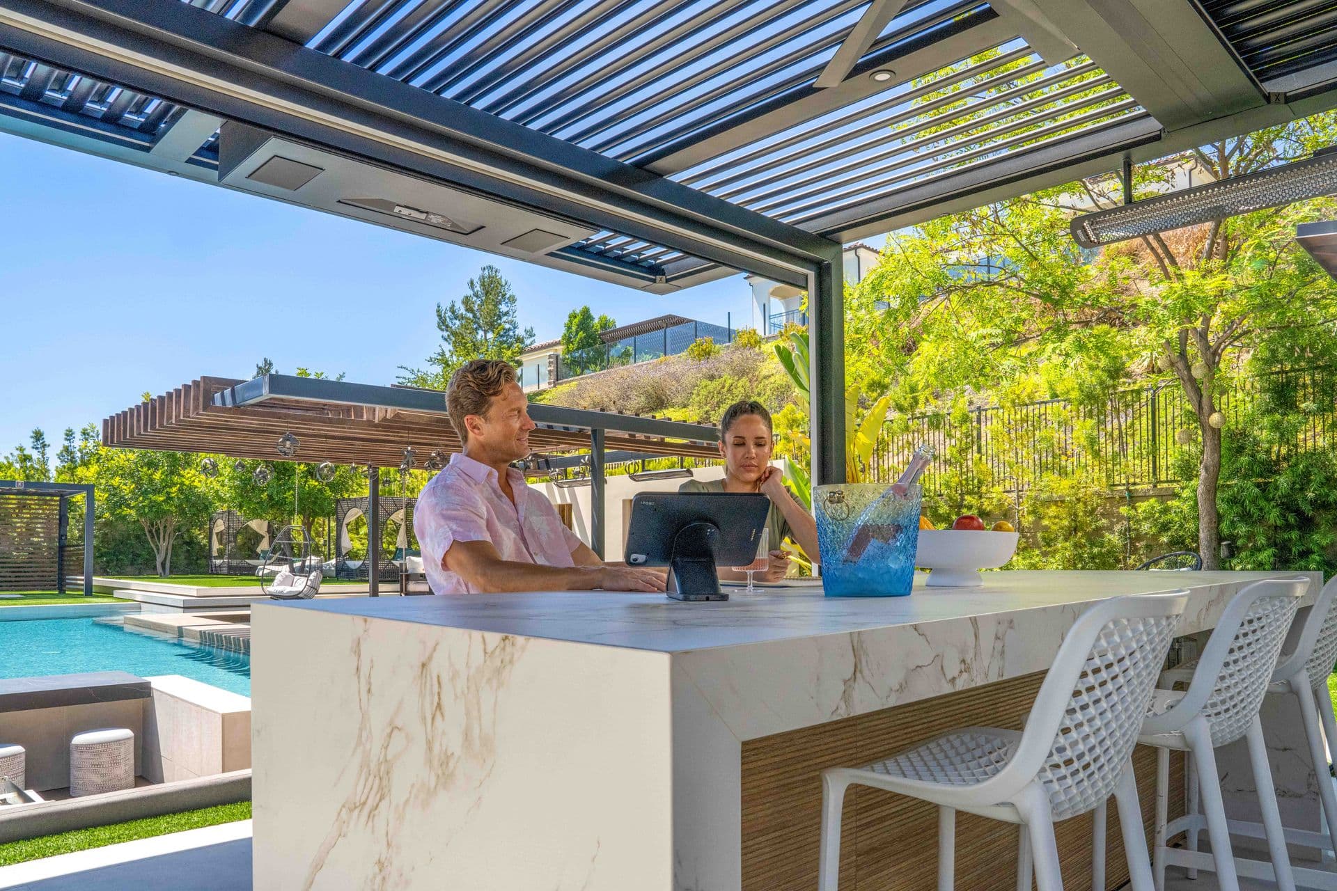A man and woman sitting under a louvered pergola by a pool with other pergolas visible in the background.