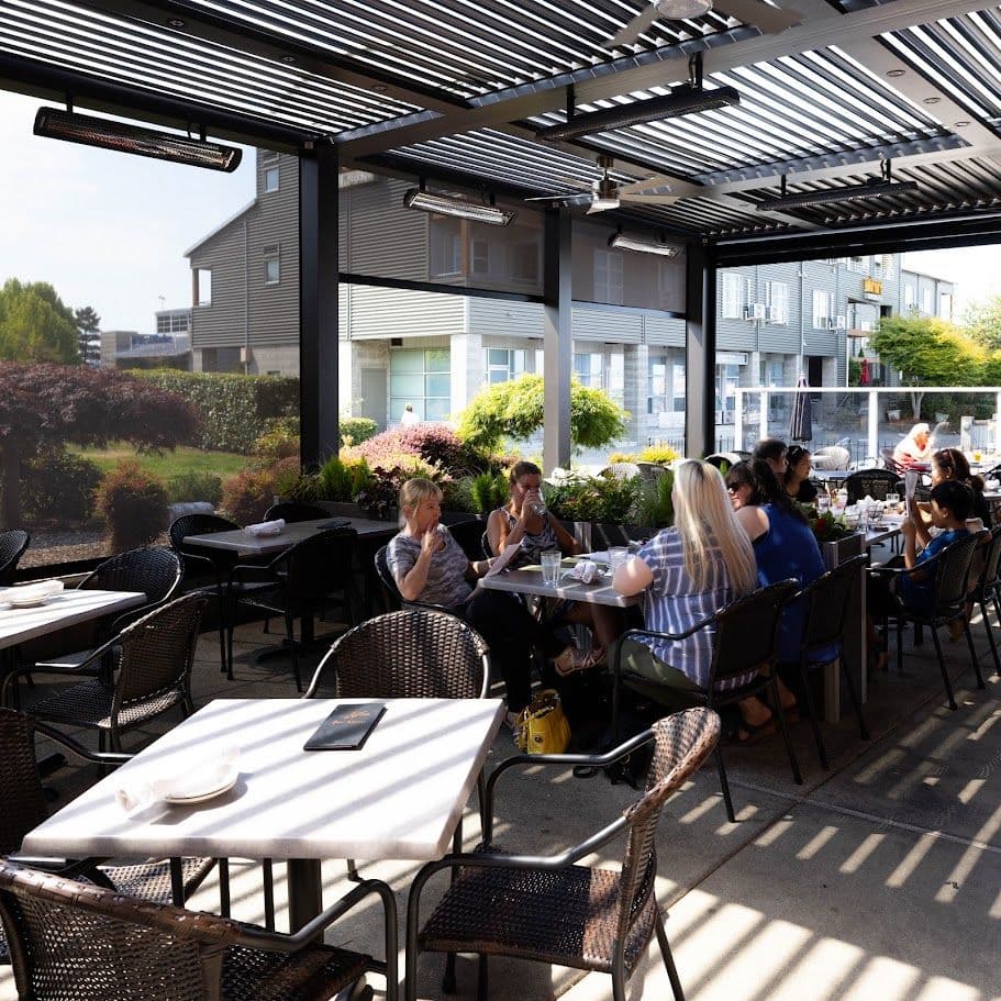 Outdoor covered patio dining area with guests seated at tables under a modern black pergola with adjustable louvered roof and heating lamps, overlooking landscaped gardens and residential buildings